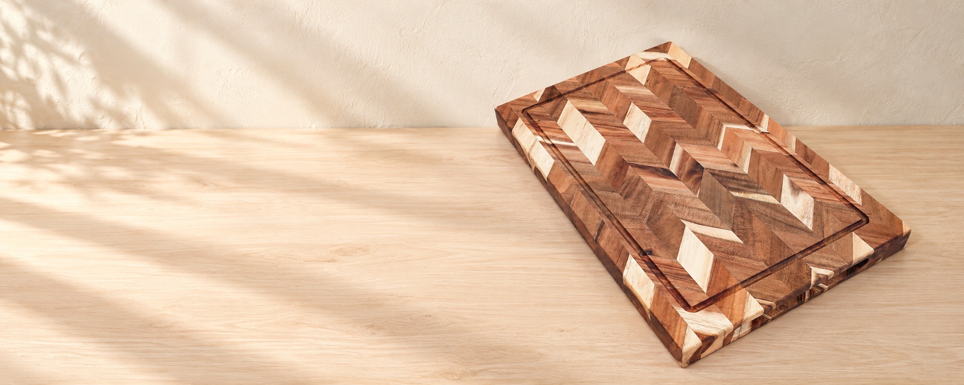 Herringbone wooden cutting board with a juice groove on a pale wooden tabletop